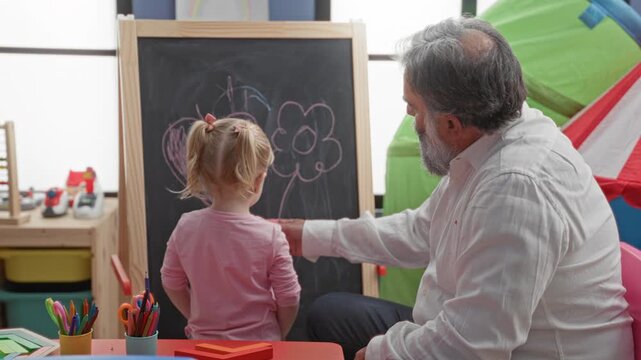 Man points finger to chalkboard while girl holds eraser and draws a flower in building; patience gentle care.