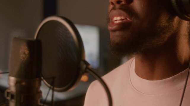 Close up view of young Black man singing into professional condenser microphone with pop filter during recording session in music studio