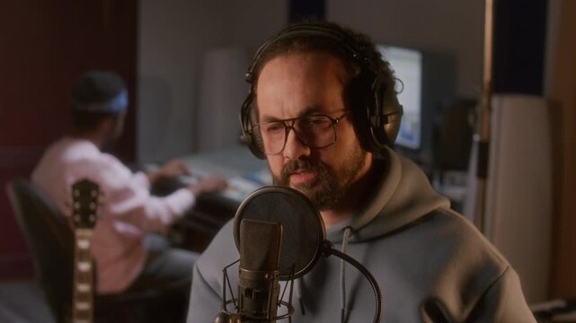 Male singer wearing headphones, hoodie and glasses performing vocal track in professional recording studio while sound technician operating mixing console in background