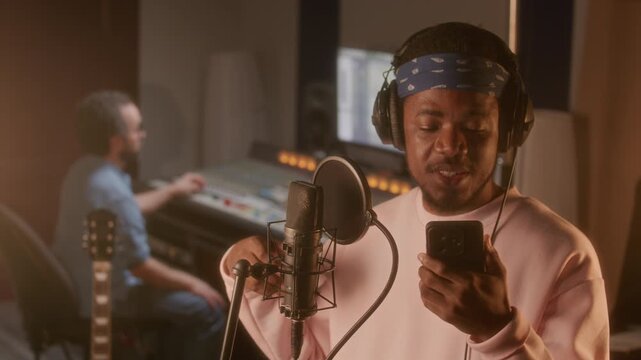 Young Black hip hop musician in headphones singing into microphone while reading lyrics from phone screen during studio recording session, sound engineer working at mixing desk in background