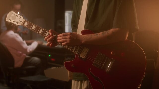 Unrecognizable male guitarist with tattooed arms playing red electric guitar during recording session in audio studio filled with warm light, sound engineers working in background. Close up shot