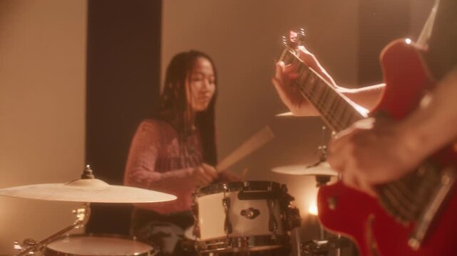 Asian female drummer playing drum set while male guitarist performing live music in foreground during rehearsal in studio filled with warm stage lighting. Rack focus