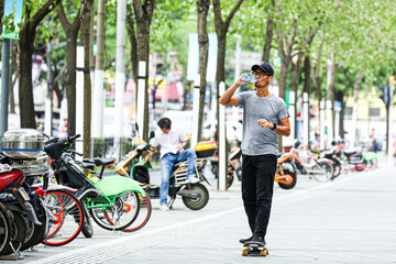 Asian man riding a skateboard and drinking water on a city sidewalk with parked scooters © Kenji