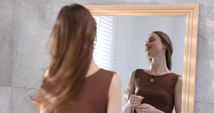 Beautiful young woman looking at herself in mirror and loosening her ponytail indoors, selective focus