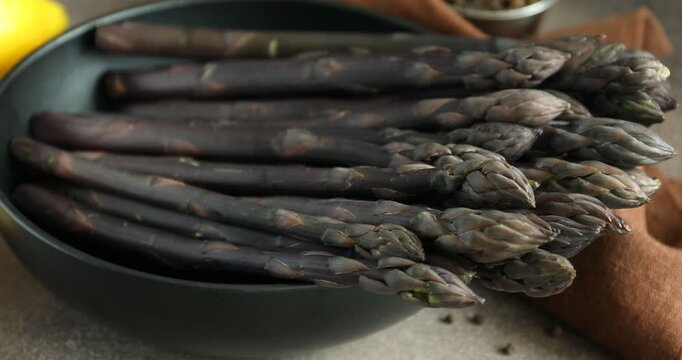 Woman putting raw purple asparagus spears into bowl at grey table, closeup
