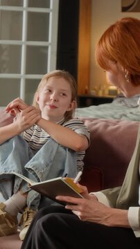 Vertical shot of smiling preteen girl talking to female psychologist feeling comfortable to express emotions while specialist note taking in journal during home visit