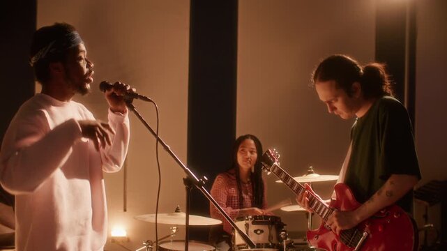 Diverse music group practicing together in warmly lit recording studio, young Black man singing in microphone while cheerful Asian girl drumming and Caucasian guitarist playing electric guitar