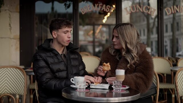 A couple is eating breakfast at a in Paris France The has a sign that says Breakfast Lunch Dinner Medium shot of the couple eating pastries and drinking coffee.