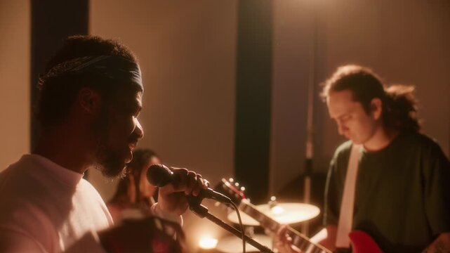Group of young multiethnic musicians practicing new song in studio: Black lead singer performing at microphone while male guitarist and female drummer providing live accompaniment in background