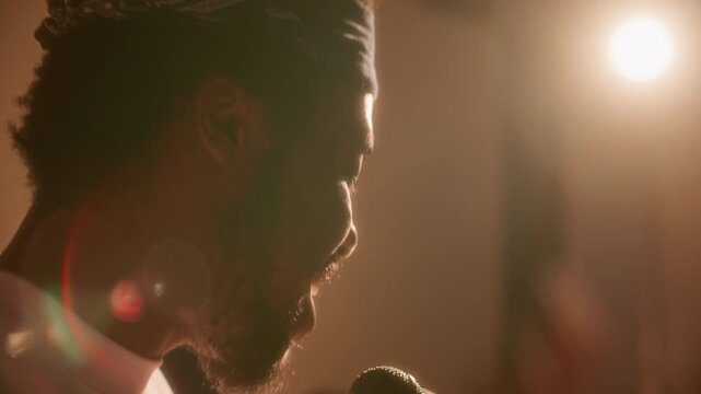Young African American hip hop singer performing emotionally at microphone during studio rehearsal, singing his lyrics and moving his hand to beat under bright stage light