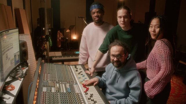 Cheerful music producer and young multiethnic band posing together for camera in professional recording studio beside audio mixing console. Group portrait