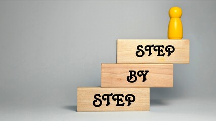 A yellow game piece stands atop a three-tiered wooden staircase labeled "STEP BY STEP," symbolizing incremental progress, goal setting, and the methodical journey toward achieving success. © FaizAhmad