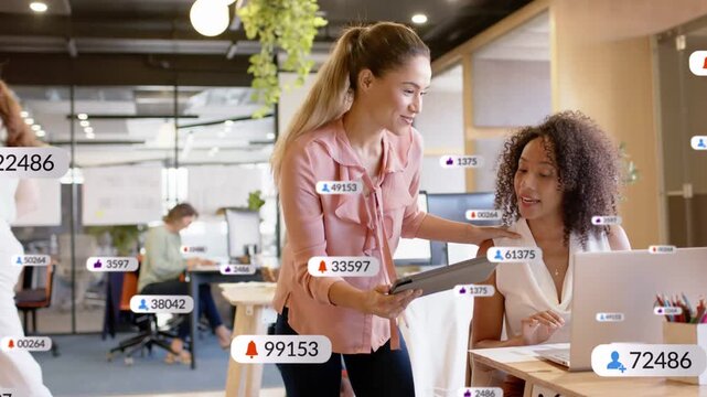 Standing woman showing tablet seated peer reviewing metrics on laptop badges rising across business