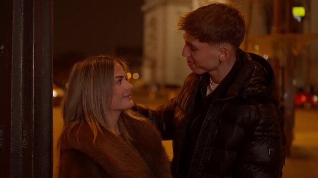 A young couple in love are talking and smiling in Paris at night near the Arc de Triomphe The cityscape is visible in the background The scene captures a romantic moment during their date in France.