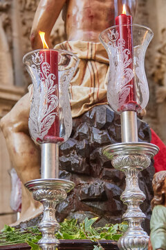 Close view of lit candles in ornate glass holders on a processional platform with a sculpture of Christ, reflecting devotion, ritual lighting and Catholic religious symbolism
