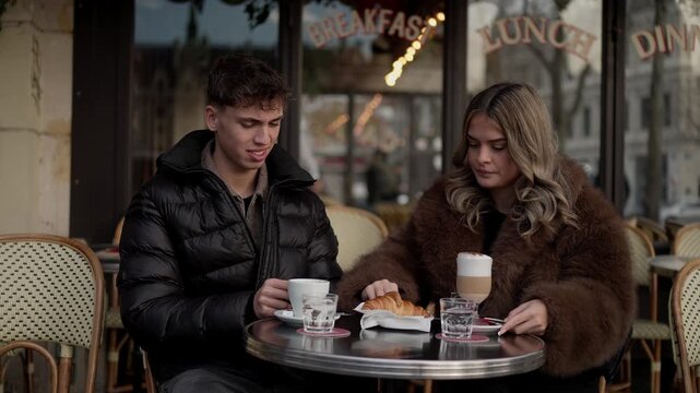 A young couple sits at an outdoor in Paris France enjoying coffee and a croissant The window displays the words Breakfast Lunch Dinner Medium shot.