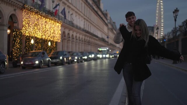 A couple walks down a street in Paris France at dusk The woman has her arms outstretched Cars and a building with lights are in the background The clip conveys a romantic travel concept.