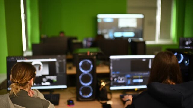 Two women engaged in creative work at computers in green-walled office, focused on screens.