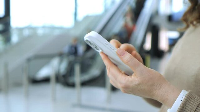 Close-up of hand holding smartphone. Person stands near escalator in modern indoor space. Background shows blurred people moving on escalator. Soft natural light fills scene. Focus remains on phone
