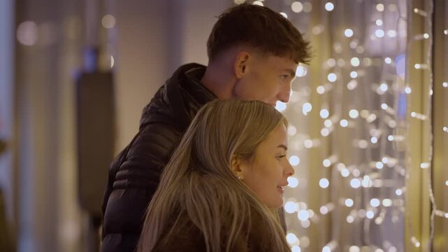 A young couple window shopping in Paris France at night The man and woman are looking at Christmas lights in a storefront display during their holiday vacation The scene captures a romantic moment in 