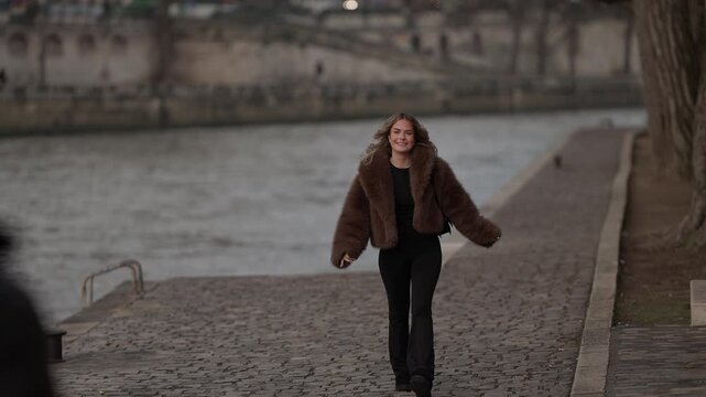 Tracking shot of a happy woman in a fur coat walking along the Seine River in Paris France The woman is smiling and enjoying the scenery on an overcast day She is strolling along the riverbank.