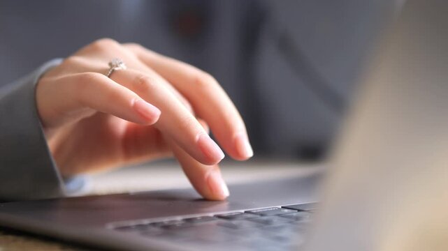 Close-up of a person's hand typing on a laptop keyboard. The hand has a ring on the ring finger. Background is softly blurred with neutral tones. Fingers move rhythmically over the keys