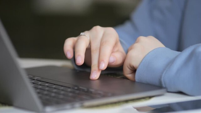 Hands type on laptop keyboard. Person wears light blue shirt. Ring visible on finger. Focused work in modern office. Laptop on desk with blurred background
