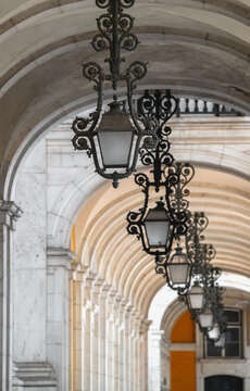 View of ornate wrought iron lanterns hanging from the vaulted stone arches of the Praca do Comercio colonnade in Lisbon, Lisbon, Portugal.