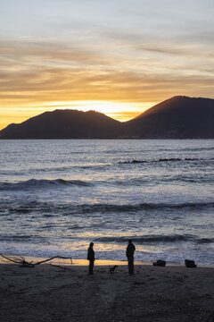 View of Venere Azzurra beach with silhouettes of two people and a dog at sunset against a backdrop of mountains and waves in Lerici, Liguria, Italy.
