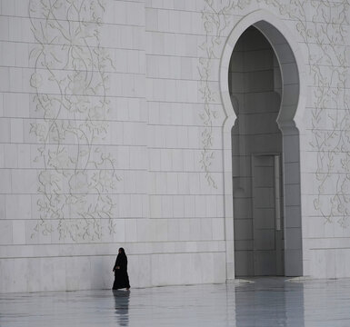 View of a woman in a black abaya walking past a white marble wall with intricate floral carvings and a large arched doorway in Abu Dhabi, Abu Dhabi, United Arab Emirates.