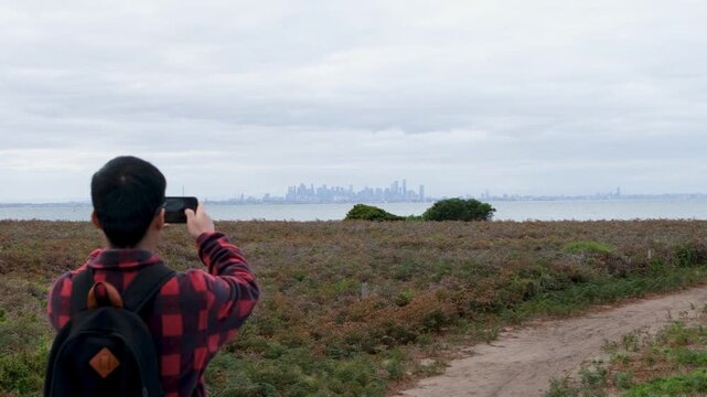 A person wearing a backpack, holding up a smartphone to capture a photo of the Melbourne city skyline in the far distance. Point Cooke Marine Sanctuary, overlooking Port Phillip Bay, Australia