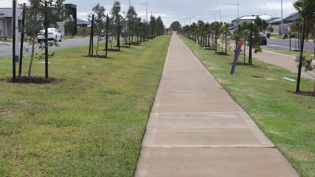 A long concrete pedestrian footpath running through a newly established residential estate in Tarneit, Melbourne, flanked by neat green nature strips and rows of young street trees secured with stakes