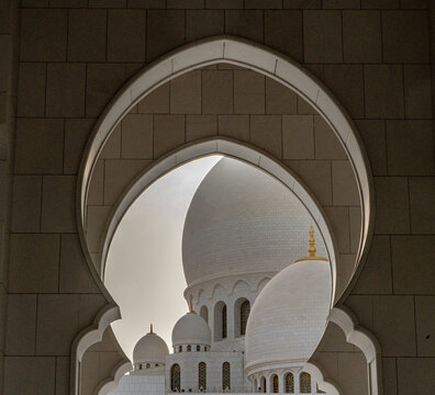 View of the white marble domes and intricate arches of a grand mosque framed by a stone archway in Abu Dhabi, Abu Dhabi, United Arab Emirates.