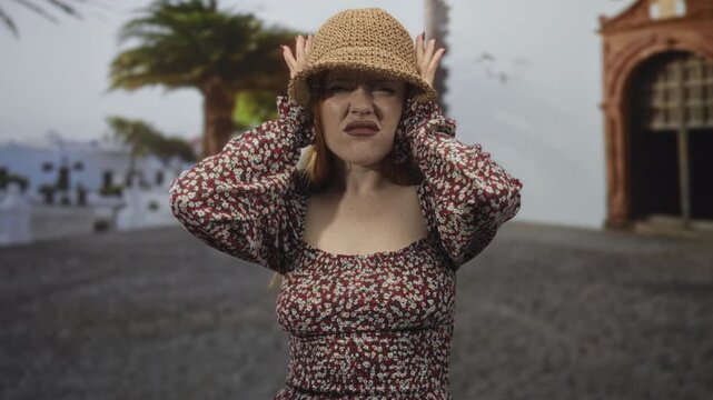 Redhead young woman holding a straw hat with both hands pressed to her head and grimacing on a sunlit street; annoyance.