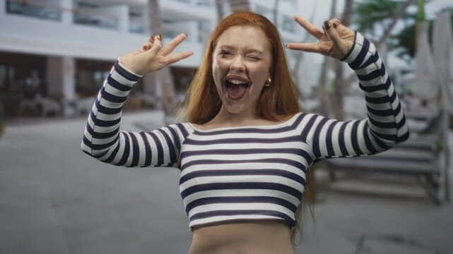 Redhead woman in striped crop top makes double peace signs, winks and sticks out tongue in a building; playful joy.