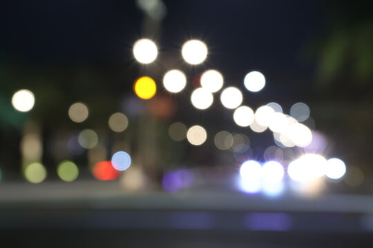 Night street glows with blurred lights and motion. Cars pass, leaving trails of brightness in the dark. Trees and signs softly fade into bokeh behind. Urban life hums under a silent, starless sky