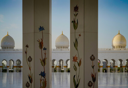 View of the ornate white marble columns with floral patterns and the majestic domes of a grand mosque under a clear sky in Abu Dhabi, Abu Dhabi, United Arab Emirates.