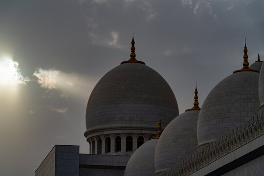 View of the white marble domes of a grand mosque with gold finials under a dramatic cloudy sky with sun rays in Abu Dhabi, Abu Dhabi, United Arab Emirates.