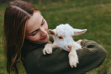 Naklejka premium Caring woman cradles a white goat kid in her arms outdoors, soft natural light, rustic woodland mood. A wholesome lifestyle moment highlighting animal bonding, tenderness, and simple joy.