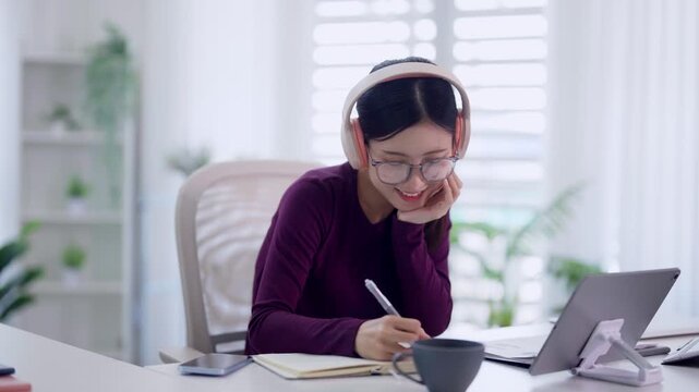 young Asian women remote working from home office wearing headphones using laptop computer taking online course writing notes notebook smiling bright modern