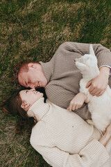 Naklejka premium Couple lying on grass, smiling, holding a white goat kid between them. Cozy sweaters and soft light create a warm, intimate lifestyle moment outdoors, highlighting connection, care, and nature.