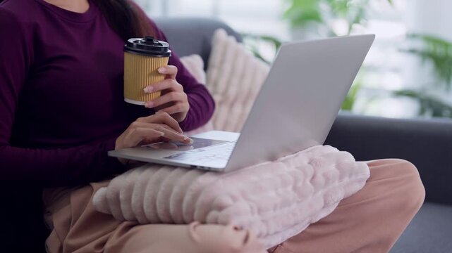 Asian woman working from home using laptop computer holding coffee cup sitting on sofa casual remote work freelance lifestyle home office comfortable cozy