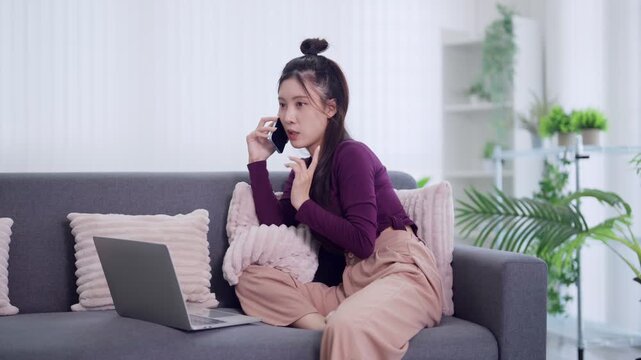 Asian woman freelancer remote work from home using laptop computer talking phone call discussing business project sitting sofa living room multitask