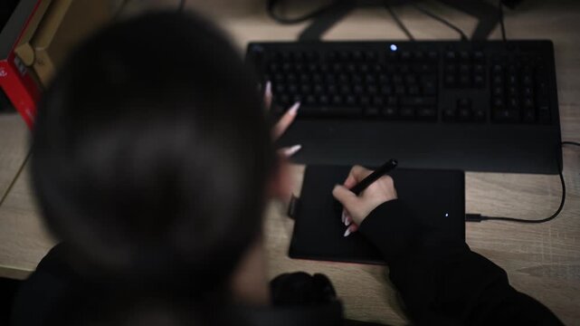 Woman drawing on digital tablet while working at computer desk in home office during night time scene.