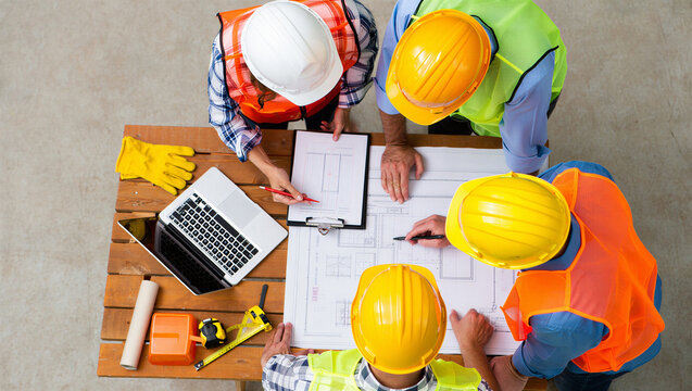 Team of four construction engineers collaborating on blueprints with laptop, top view.