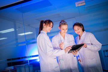Three students in lab coats work together on a tablet in a science lab during evening hours in a modern research facility