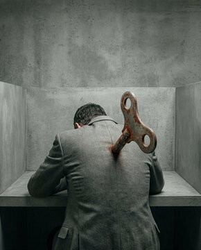 Exhausted businessman with large rusty wind-up key in his back slumping over desk in minimalist grey office environment