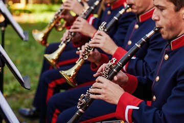 Fototapeta premium Orchestra band members play clarinets outdoors wearing blue uniforms with red trim. Musicians perform with sheet music stands for a concert. Brass instruments are visible in background.