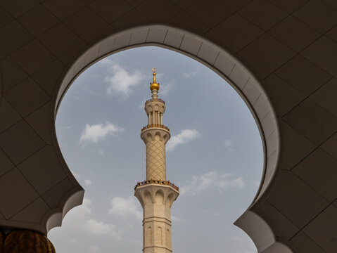 View of a tall ornate minaret with a golden crescent moon on top framed by a white scalloped archway under a blue sky in Dubai, Dubai, United Arab Emirates.