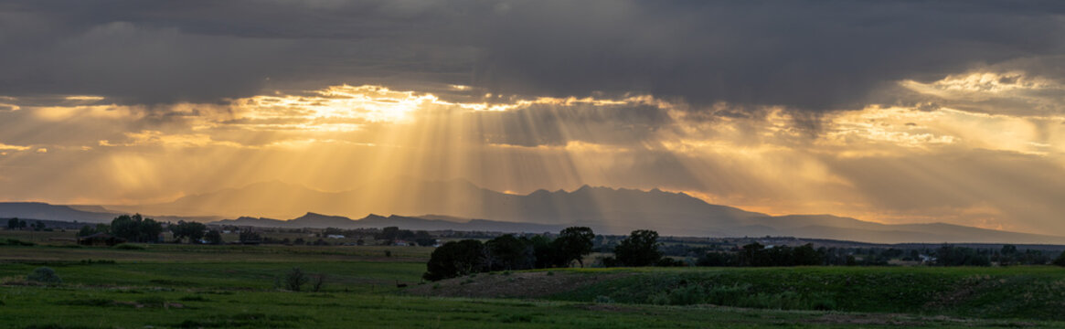 View of dramatic sun rays breaking through dark storm clouds over a mountain range and green fields at sunset United States.